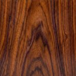 A close-up view of a polished Santos Palisander surface, displaying a warm spectrum of reddish-browns and oranges intersected by distinct, dark brown grain lines.